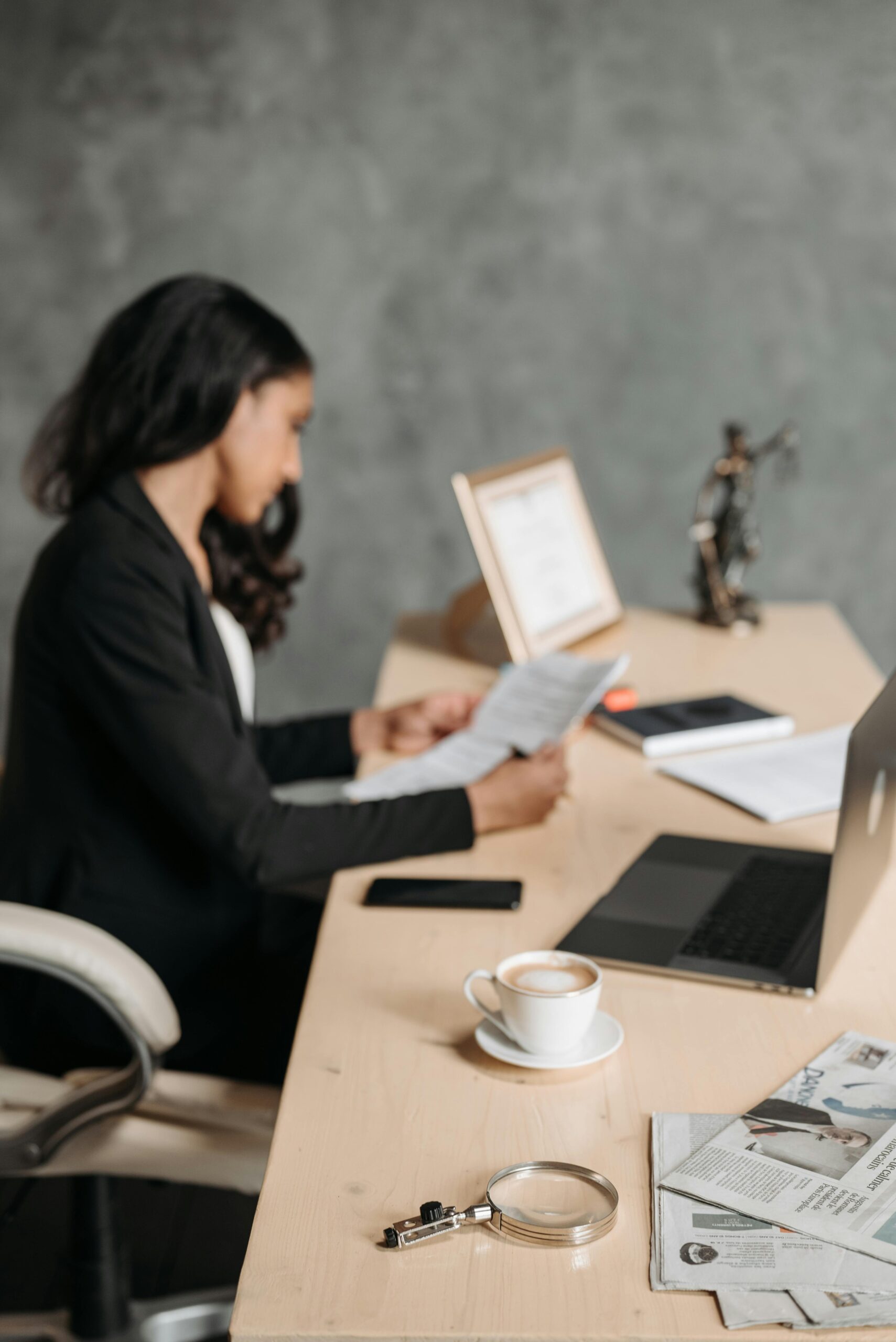 Women dressed in professional attire working at her desk reading documents with an open laptop, coffee mug and a phone on the desk.
